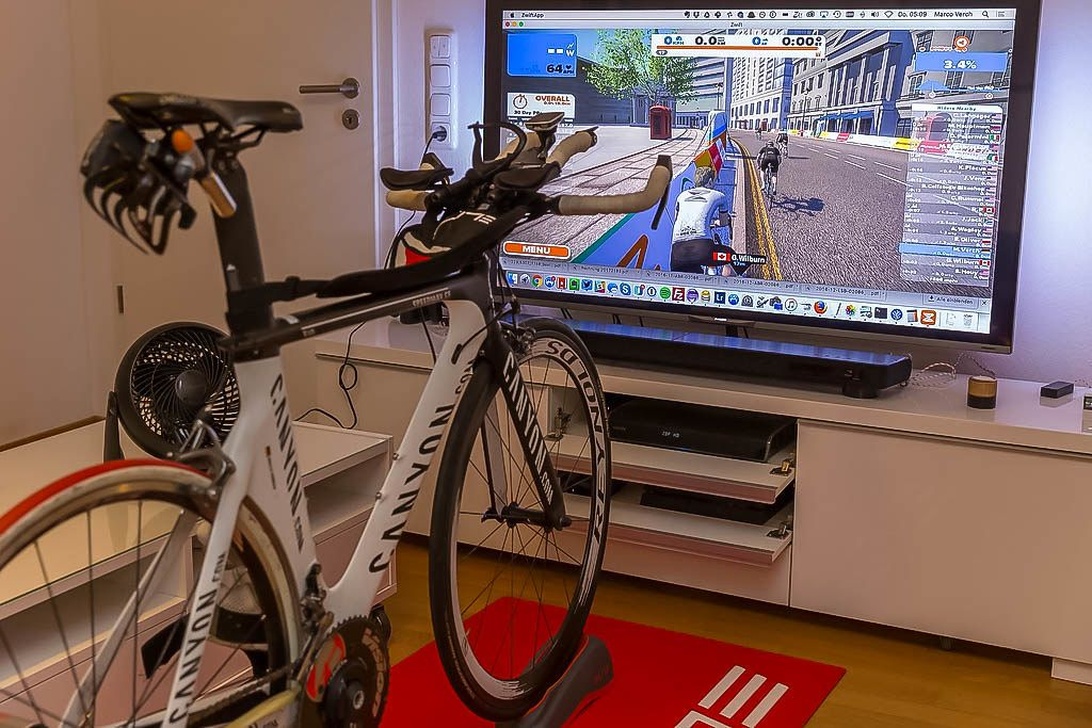 En inomhuscykeluppställning med en racercykel monterad på en trainer framför en TV som visar ett virtuellt cykelprogram.